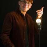 portrait d'un homme joyeux en casquette marquée Craft et en chemise de velours avec une ampoule allumée